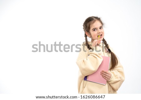 Attractive young girl student with pigtails dressed in beige hoodie holds in hands notebook and pen in mouth, looking at the camera, isolated over white background, copyspace, studio photo