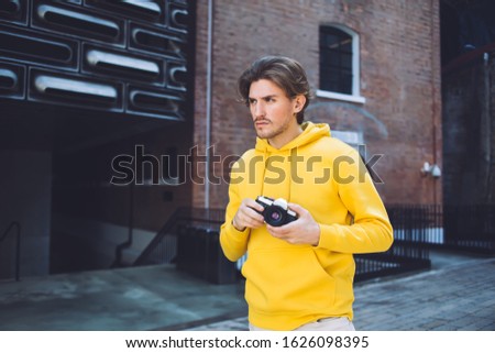 Handsome stylish serious thoughtful young male wearing yellow sweatshirt holding photo camera while walking on street with brick building on background