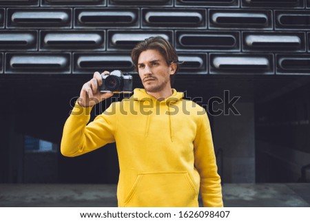 Handsome hipster man wearing bright yellow hoodie standing with back to covered parking entrance holding photo camera in raised hand and choosing object for next picture