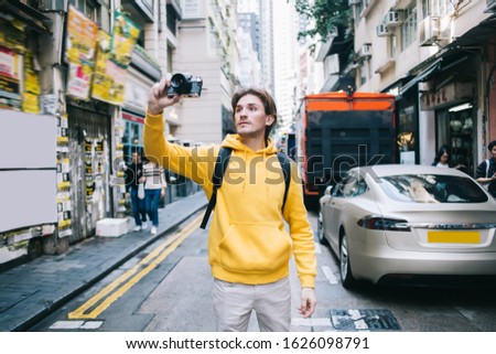Young male traveller with backpack spending vacation in city walking along street and taking photo with camera exited by local views