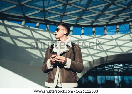 Low angle of handsome young male teenager in coat and sunglasses standing near shopping center with smartphone and looking away