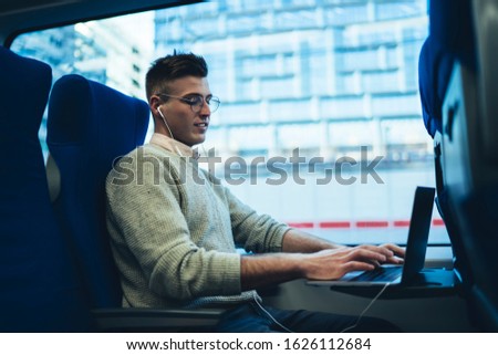 Side view of young handsome male freelancer in sweater and glasses using laptop and earphones while traveling by bus on background of street in window