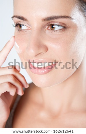 Close up detail view of an attractive young woman applying hydrating care cream on her face with her hand and finger, while smiling isolated against a white background, indoors.