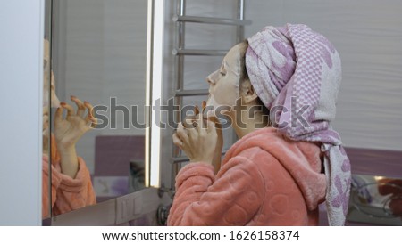 Woman applying cosmetic face mask and looking at mirror in bathroom. Girl taking care of her face skin. Skincare spa treatment. Facial mask