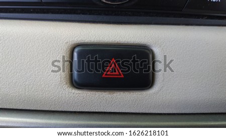 A car's hazard lamp button, red triangle in black colored button surrounded by white / beige colored dashboard