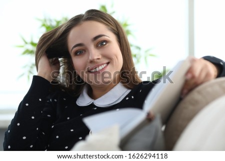 Portrait of happy young female smiling on camera and touching hair. Lovely relaxed lady sitting on sofa with book indoors. Spare time and hobby concept