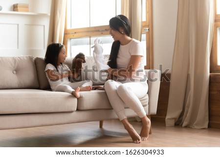Happy Asian young mom sit on couch playing with cute little ethnic daughter in living room, overjoyed Vietnamese millennial mother or nanny have fun engaged in funny game with small girl child