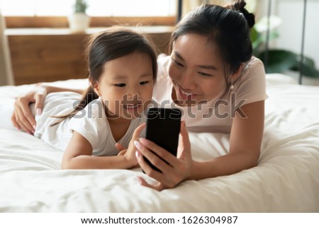 Happy Asian young mom lying in bed with cute small biracial daughter watch video on modern smartphone together, smiling Vietnamese mother relax in bedroom with little girl child use cell at home