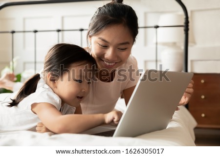 Happy young Asian mom and little ethnic daughter lying in bed at home watch video on modern laptop, smiling Vietnamese mother or nanny relax in bedroom with small biracial girl child using computer