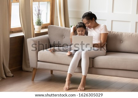 Happy young Asian mother relax on couch with cute small biracial daughter watch video on laptop together, smiling millennial Vietnamese mom rest on sofa use computer gadget with little girl child
