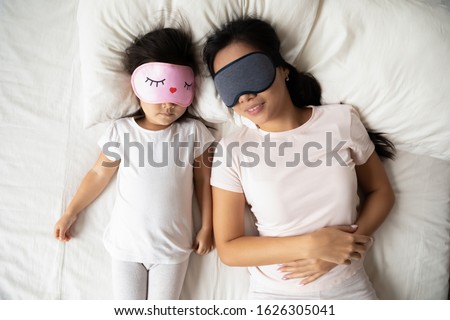 Top view of calm Asian mom and daughter in sleeping mask lie relax in comfortable white bed together, Vietnamese young mother and cute little girl child rest take nap daydream in cozy home bedroom Royalty-Free Stock Photo #1626305041