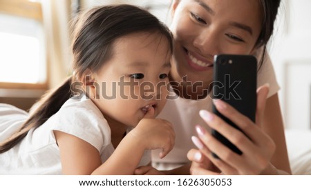 Smiling Asian millennial mom or nanny relax with little biracial girl child using cellphone together, happy Vietnamese young mother rest with small ethnic daughter watch video learning on smartphone Royalty-Free Stock Photo #1626305053