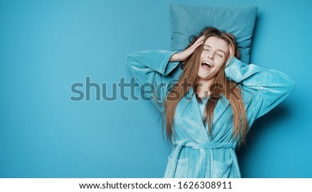 Waking yawning long haired young lady in dressing gown with her head on pillow over isolated background, prevailing blue tone Royalty-Free Stock Photo #1626308911