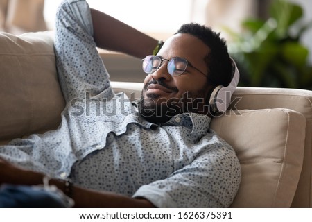 Serene satisfied young adult african american man lying on comfortable sofa wear wireless headphones enjoy listen modern music audio book with eyes closed meditate relax feel no stress chill at home Royalty-Free Stock Photo #1626375391