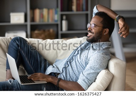 Relaxed millennial african man resting on couch using laptop at home, smiling young afro american hipster guy lounge on sofa enjoy leisure time with technology device looking dreaming feel satisfied Royalty-Free Stock Photo #1626375427