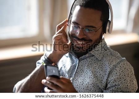 Smiling young adult african man wearing wireless headphones listening mobile music on smartphone, happy afro american guy using online player podcast audio book in app on cell phone relaxing at home
