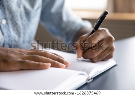 Close up view of african left-handed businessman writing in notebook, american male hands holding pen making notes planning new appointments information in organizer personal paper planner at desk Royalty-Free Stock Photo #1626375469