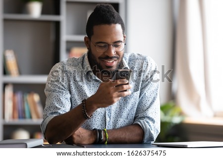 Happy young african business man student professional user hold smartphone modern gadget texting message in social media app online sit at home office desk using cell phone technology for surfing web