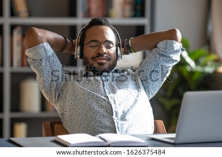 Relaxed african business man wear wireless headphones enjoy listen music with eyes closed hand behind head sit at work desk, young worker take break in office feel peace of mind stress relief concept Royalty-Free Stock Photo #1626375484