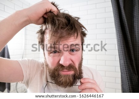 Overgrown man, with a shaggy hairstyle and beard, looks in his reflection in the mirror and is dissatisfied with his appearance.
