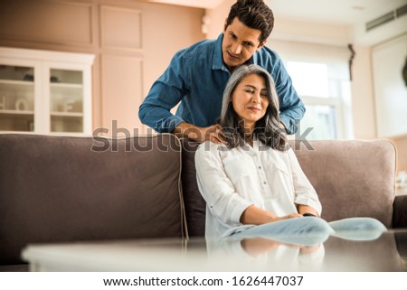 Handsome adult husband standing near sofa while doing massage for his pretty wife stock photo