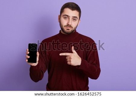 Picture of serious man dresses dark burgundy sweater looking directly at camera while holding smartphone and pointing at him isolated over lilac background, bearded guy with smart phone in hands.