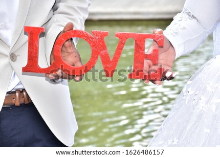 Newlyweds, the bride and groom in white hold in their hands the word love from scarlet letters in English. Against the background of green lake water, together, background, invitation, greeting card, 