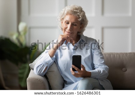 Happy older woman sitting on comfortable couch, looking at smartphone screen. Smiling hoary 60s grandmother making selfie shot, using mobile apps, watching video or calling grown up children at home.