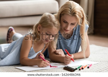 Young blonde woman nanny babysitter lying on floor carpet with cute small kid girl, drawing coloring pictures in albums. Happy mother enjoying creative activity with little preschool daughter.