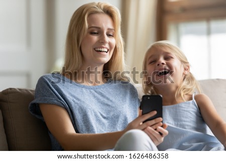 Happy young blonde attractive mother showing funny cartoon on smartphone to laughing lovely cute little preschool daughter, relaxing together in living room. Adorable family of two having fun at home.