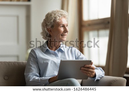 Smiling elderly mature woman sitting on cozy couch, holding computer tablet, looking away at window. Happy dreamy middle aged grandmother thinking of future, planning trip, enjoying free time.