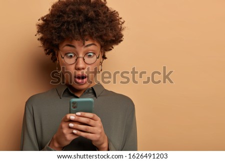 Horizontal shot of shocked female blogger watches video on smarphone with amazed face, gasps from wonder, surprised about message, reads unbelievable news, wears round spectacles and grey shirt