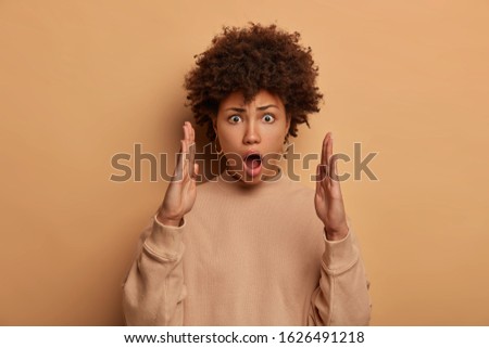 Embarrassed dark skinned woman shapes huge object with both palms, shapes size of big box, keeps mouth opened and eyes popped out, wears casual jumper, explains width of something, exlaims loudly Royalty-Free Stock Photo #1626491218