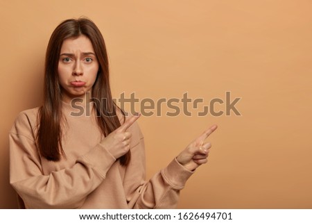 Indoor shot of unhappy disappointed woman points on blank space, missed opportunity, asks your opinion, makes perplexed choice, purses lower lip, isolated over brown background, expresses regret Royalty-Free Stock Photo #1626494701