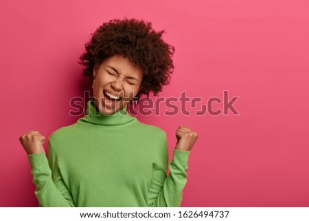 Great achievement and success concept. Happy Afro American woman clenches fists, makes victory gesture, screams yeah, tilts head, wears green turtleneck, celebrates achieving goal, isolated on pink