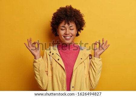 Concentrated cheerful curly woman keeps both hands in okay gesture, meditates indoor, has eyes closed, wears yellow anorak, smiles gently, stands against vivid background. Body language concept