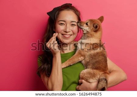 Attractive brunette woman with eastern appearance, holds Shiba Inu dog on hands, makes korean like sign, expresses love to pet, adopts animal, smiles broadly, going to groomer or outdoor walk