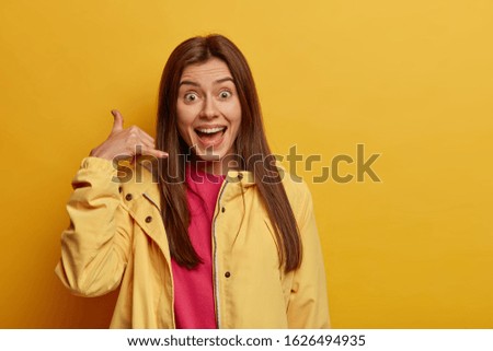 Body language concept. Positive brunette woman makes call gesture, says call me back, wears yellow anorak, asks for number, looks gladfully at camera, isolated over vivid background, copy space area