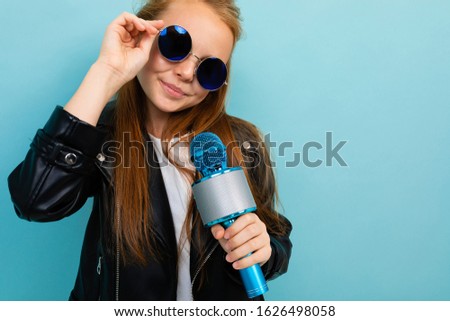 Caucasian teenager girl with brown hair in black jacket, blue sunglasses with blue microphone isolated on blue background