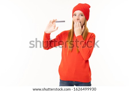 Happy teenager girl with red hair, red hoody and hat with credit card isolated on white background