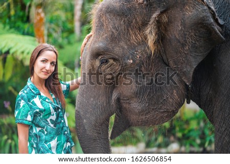 A Caucasian woman in a bright shirt poses next to an elephant. Close up. Jungle forest on the background