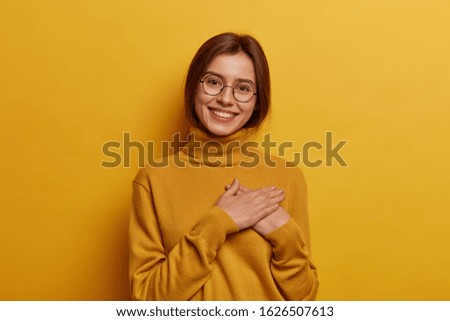 Glad European girl feels pleased and grateful, being thankful for awesome gift, presses hands on chest, has pleasant tender smile, tilts head, wears yellow turtleneck. What pleasure to meet you Royalty-Free Stock Photo #1626507613