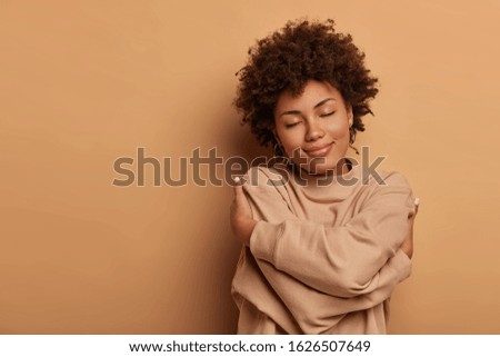 Love yourself, self acceptance. Gentle lovely Afro American woman crosses hands and embraces own body, tilts head and closes eyes, wears comforable brown sweater, models indoor. I love myself Royalty-Free Stock Photo #1626507649