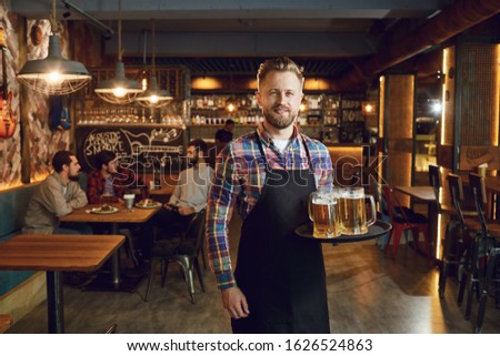 Bearded waiter with a tray of glasses of beer against the background of a pub bar Royalty-Free Stock Photo #1626524863