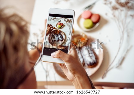 Cropped view of young adult blogger taking food photography on her modern smartphone. Top view of woman holding device cover plate with dessert and making shot