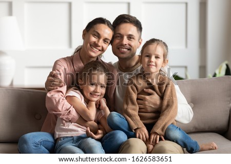 Portrait of loving young parents sit on couch in living room hug little daughters look at camera, happy family with girls kids rest on sofa posing for picture at home together, show unity and support