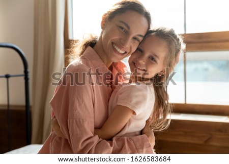 Portrait of happy young mother hug cuddle with little preschooler daughter relaxing in home bedroom together, smiling mum or nanny embrace look at camera with small girl child show love and care