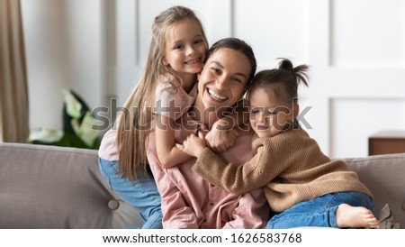 Portrait of overjoyed young mother sit on couch in living room cuddle with little preschooler daughters, happy loving mum pose look at camera relax at home enjoy family weekend with small girl kids Royalty-Free Stock Photo #1626583768