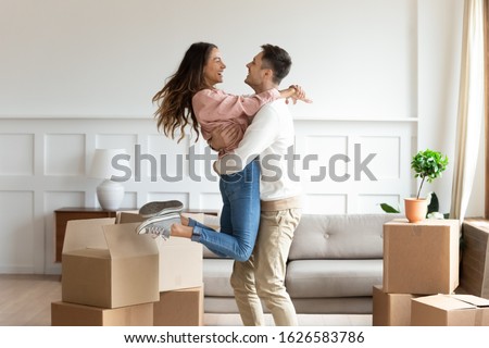Overjoyed Caucasian husband hold carry swirl happy wife excited to move in new home together, smiling young couple dancing in living room have fun on relocation day in apartment, ownership concept
