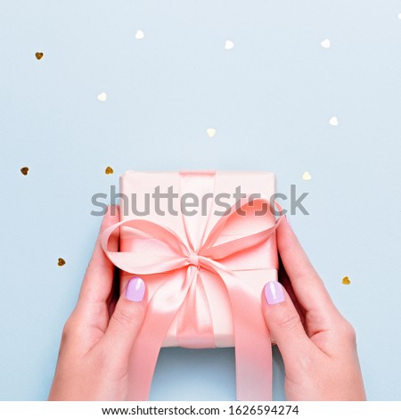Woman hands holding gift box in pink color on pastel blue background with heart shape confetti, copyspace. Valentine's Day greeting card in trendy colors, Happy Valentine Day background